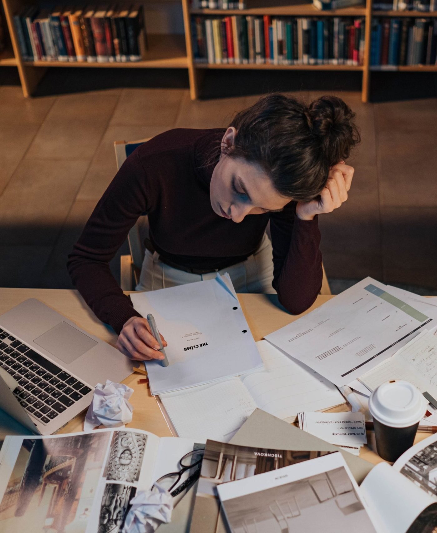 Woman studying at desk with documents, laptop, and coffee in a cozy library setting.